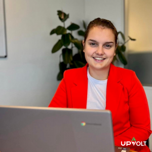 Een jonge vrouw zit aan een bureau en werkt op een laptop. Ze draagt een rood jasje en heeft een glimlach op haar gezicht. Achter haar staat een plant. In de rechterbenedenhoek van de afbeelding staat het logo van Upvolt.